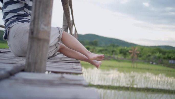 慢动作年轻女性坐下来的腿挂在木桥的边缘，放松摆动的腿，亚洲乡村边文化传统的稻田，平静的放松，下半身段
