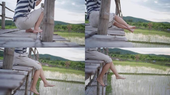 慢动作年轻女性坐下来的腿挂在木桥的边缘，放松摆动的腿，亚洲乡村边文化传统的稻田，平静的放松，下半身段