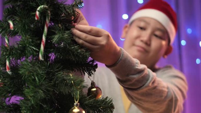 亚洲男孩戴着圣诞老人的帽子，在家里的圣诞树上悬挂装饰品，准备概念。
