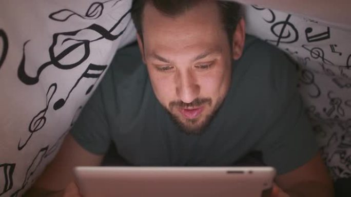 一个留着胡须和胡须的年轻男人的脸特写，在毯子下使用数字平板电脑，晚上在床上聊天和挥手