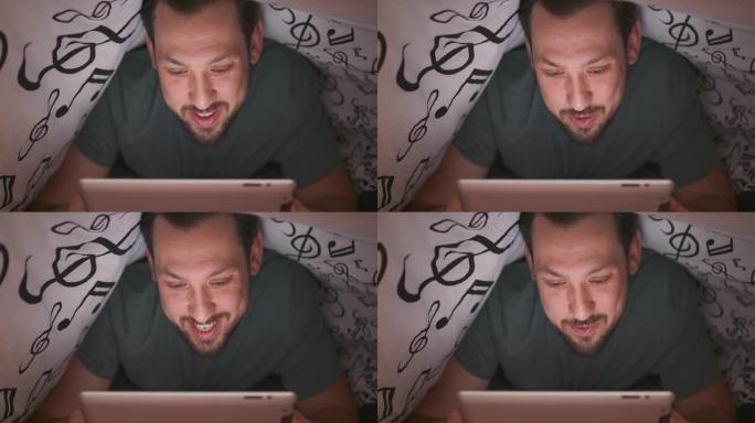 一个留着胡须和胡须的年轻男人的脸特写，在毯子下使用数字平板电脑，晚上在床上聊天和挥手