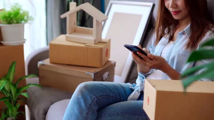 在搬家的日子里，快乐的年轻亚洲妇女在新房子的客厅里使用智能手机，里面放着一堆纸板箱