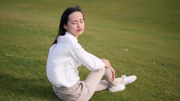 穿着白衬衫的美丽中国年轻女子的肖像坐在绿草地上，闭着眼睛，享受微风吹着她的长发，睁开眼睛，最后微笑着
