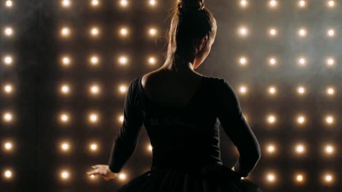 穿着黑色连衣裙的芭蕾舞演员的剪影是在黑暗的工作室里跳芭蕾舞。
