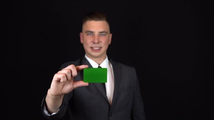 一个穿着西装的年轻商人手里拿着一张银行卡。孤立的黑色背景。Chromakey绿卡。