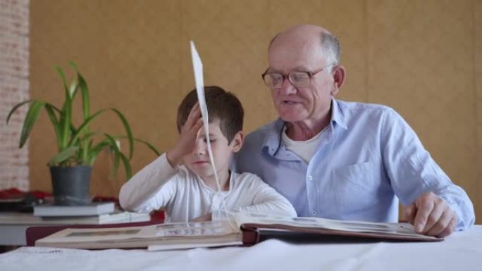 戴着眼镜的老人和他的孙子一起欣赏快乐的回忆，看着一张家庭相册坐在房间的桌子旁