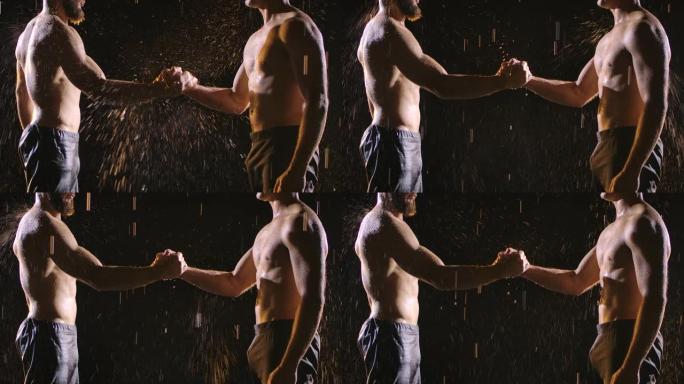 两个肌肉发达的赤裸上身的男人在飞溅的雨中握手。在黑色背景的工作室里拍摄的照片。慢动作。特写