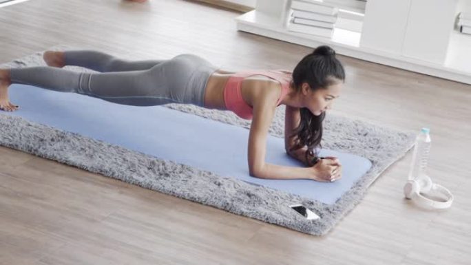 穿着运动服的美丽亚洲女性在家里的客厅做木板运动瑜伽锻炼。