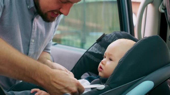 车内摄像头。特写男婴坐在车内的婴儿汽车座椅上。年轻的大胡子父亲检查婴儿系好的安全带，并与他交谈。慢动