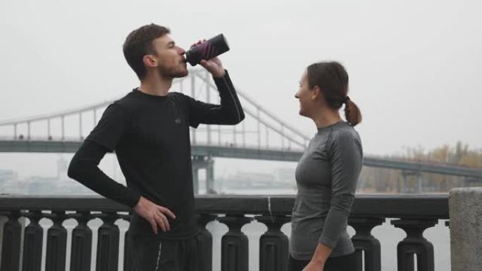 穿着运动服的快乐可爱运动活跃的夫妇在河边跑步训练和饮用瓶子里的水后休息和放松。职业男女选手谈笑风生