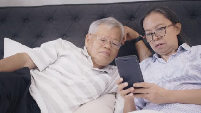 亚洲老年夫妇在周末假期在家中卧室背景下使用智能手机阅读社交媒体。老年人幸福生活的概念。4k慢动作。