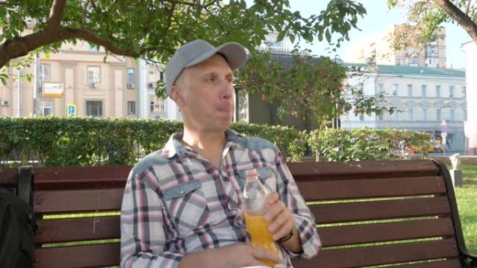 一个白人男子坐在公园的长凳上喝果汁四处张望