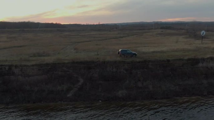 越野黑色suv汽车在金色日落景观的河岸上行驶。在傍晚的阳光下，跨界车沿着海水在海岸上行驶。