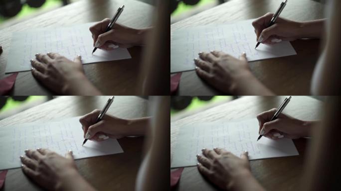 年轻女子用笔在白纸上写给她心爱的新郎的情书，坐在阳台附近房间的桌子旁。阳台外的绿草。婚礼准备。新娘的