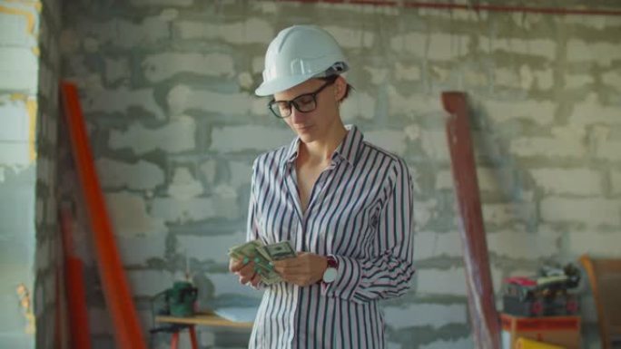 年轻微笑的女工戴着白色安全帽数钱，站在建筑工地。工头持有现金用于房屋装修过程。“热固性粉末混合” 盒