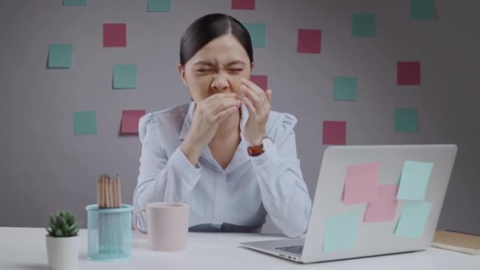 在笔记本电脑上工作的亚洲妇女生病了，发热打喷嚏，坐在家庭办公室。