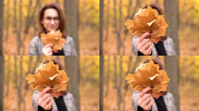 一位戴着眼镜的年轻女子躲在秋天森林中的一棵大叶树后面。女孩对着镜头伸出树叶。