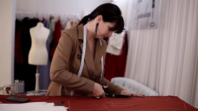穿着时尚西装和耳环的女性时装设计师在工作室里穿着红色面料，里面装满了裁缝工具-图案，剪刀，卷尺。女裁