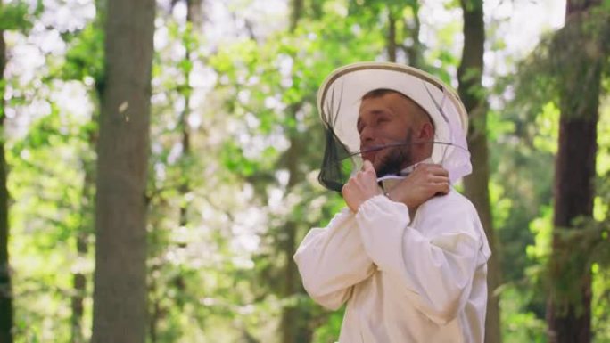 大胡子的年轻英俊的养蜂人系上白色防护服的拉链，戴着圆形的防护罩，微微微笑，呆在森林的树木之间。背景中