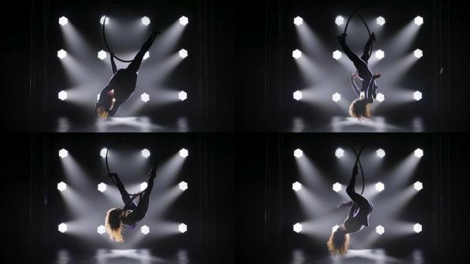 美丽的体操运动员倒挂在空中的铁环上，表演杂技元素。穿着紧身紧身衣的苗条身材的轮廓。在有舞台灯光的黑暗