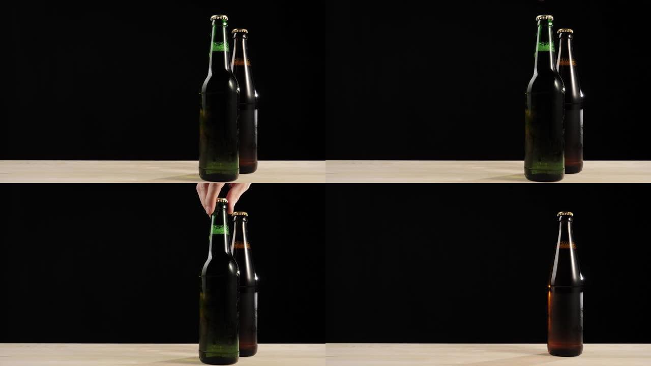 新鲜啤酒。手从黑色背景下的木制桌子上拿了一个绿色的瓶子，里面有美味的工艺啤酒，靠近棕色瓶子。水滴冷新