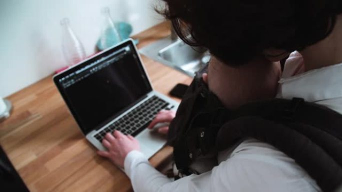 母亲在笔记本电脑键盘上打字，婴儿睡在婴儿背带中