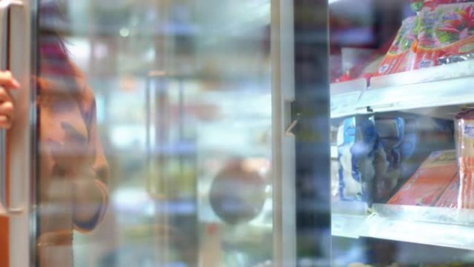 在COVID-19大流行期间，年轻的亚洲女性戴着口罩在超市购物，看着货架冰柜。女性买家选择产品 在一