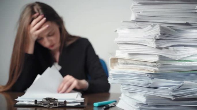 办公室办公桌上堆放着一堆未完成的文件和文件。过度工作的女性办公室会计坐在文件前，握着她的头与手，疲惫