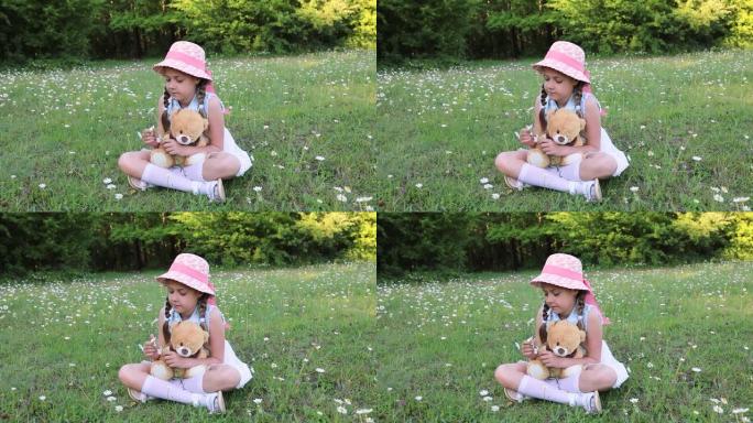 一个戴着帽子的可爱的小女孩在玩她的泰迪熊。婴儿和最喜欢的玩具。夏天，绿色林间空地