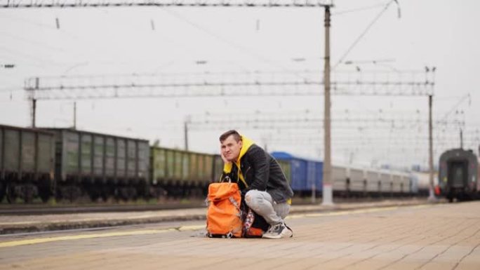年轻人蹲在站台上，等待火车。男性乘客，背包坐在铁路平台上等待火车。旅游、旅行和娱乐的概念