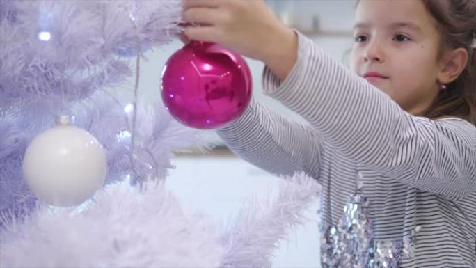 漂亮的小女孩从模糊的背景来到最前面的圣诞树上，把一个玻璃美女放在树枝上。