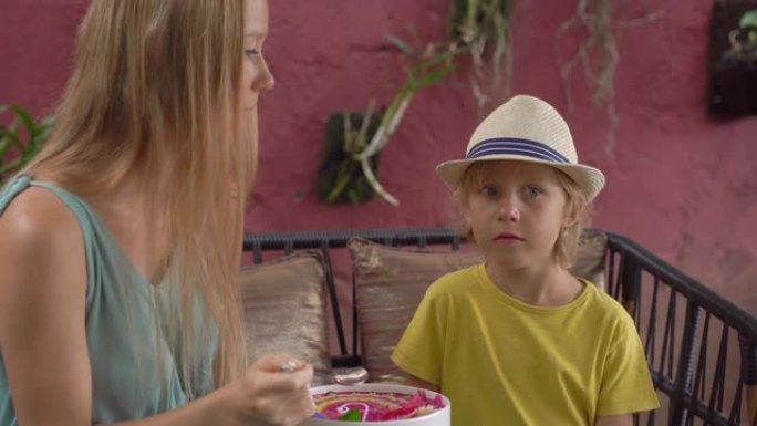 年轻女子和她的小儿子坐在一个美丽的咖啡馆里，在碗里享受五颜六色的格兰诺拉麦片冰沙。健康饮食理念