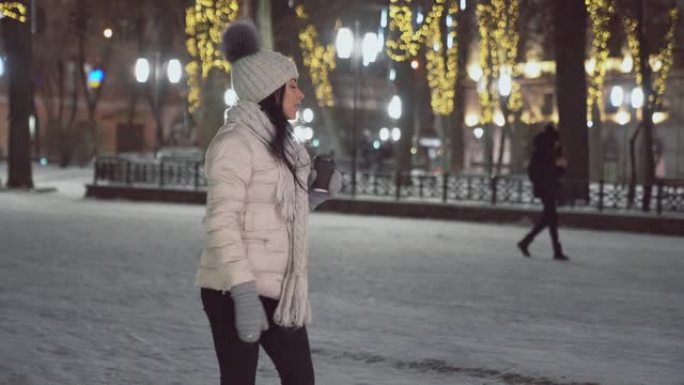 穿着粉红色夹克的英俊女孩，带pom pom的羊毛帽子，skarf和手套在晚上在城市散步。街上下雪的年