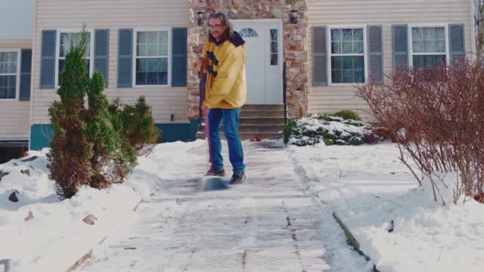 一名身穿黄色夹克的50岁成年长发男子扫过小路，在一场冬季降雪后，将乡间别墅前院的积雪从积雪中扫走。美