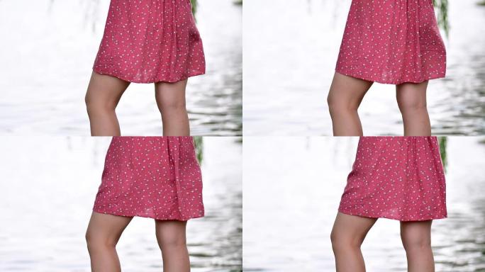 红色连衣裙随风飘扬，模糊的湖背景，一部分成年年轻女士站在公园的湖边，享受微风吹着她的连衣裙，超级慢动
