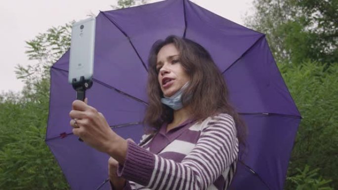 一个戴着冠状病毒面具的年轻白人妇女正在森林里伐木。在森林中，一个拿着雨伞的雨伞下的女人使用智能手机进
