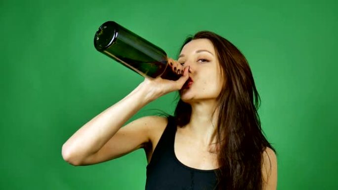 可爱的喝醉的长发女孩正在工作室里喝绿色背景下的黑瓶葡萄酒