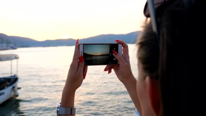 女人在智能手机上用金色的日落拍摄风景优美的海洋景观。女孩用手机得到美丽的海景照片。晚上享受在海边休息