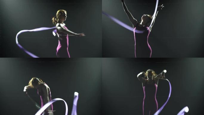 身材紧身的苗条年轻女体操运动员的轮廓，用丝带做体操运动。聚光灯下的黑色烟熏背景。特写
