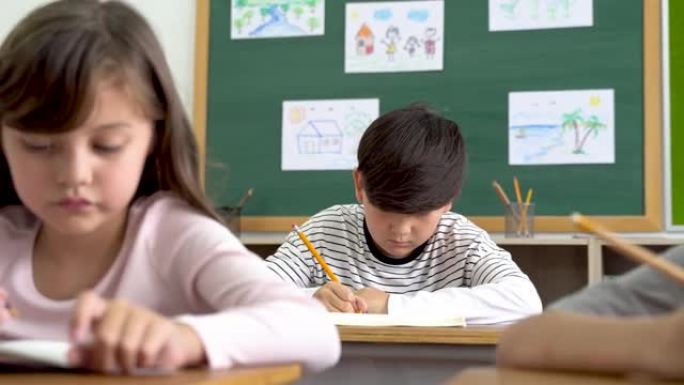 快乐的女学生坐在教室的桌子上微笑的肖像。可爱的高加索小学女孩坐在学校在纸上写字，看着相机，教育，学习