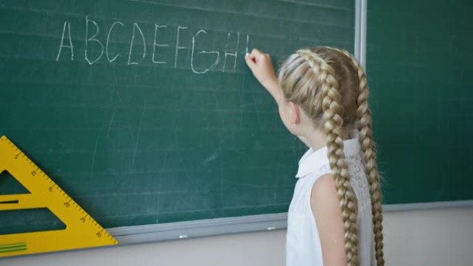 黑板上快乐的女学生的肖像在教室里用白色粉笔写字，看着相机，小学英语课