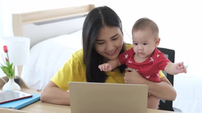 美丽的商业亚洲妈妈正在使用笔记本电脑，微笑着与她可爱的宝宝在一起。休产假的年轻母亲试图带着蹒跚学步的