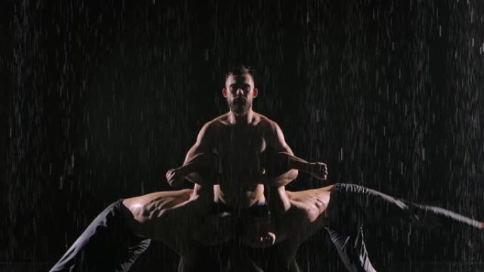 一组三个杂技演员在雨中表演困难的平衡技巧。男人以慢动作站在黑色背光背景下。特写