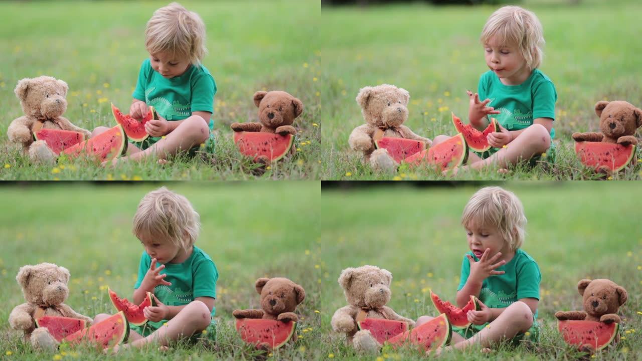 可爱的蹒跚学步的小孩，金发男孩，和一些泰迪熊朋友在公园里吃西瓜
