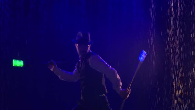 一个时髦的男人在蓝光照亮的水滴背景下表演的灯光表演。艺术家在晚上被荧光棒剪影。慢动作。特写