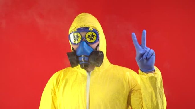 一个穿着黄色防护服和戴着防护面具的男人露出两个手指，一个和平标志。防护概念，放射性危害概念。红色背景
