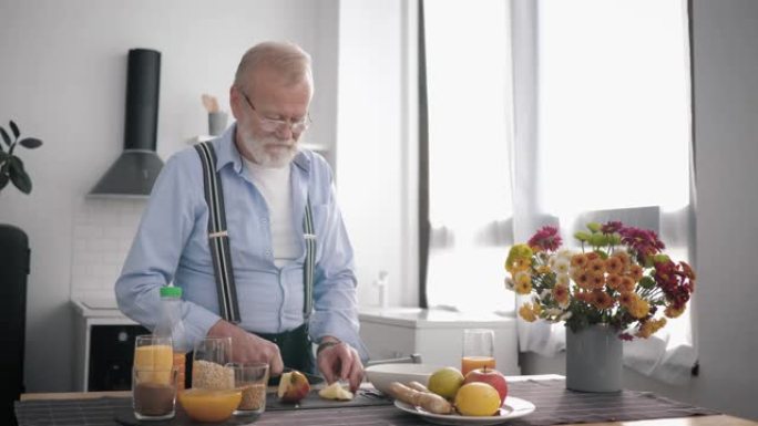 戴眼镜胡子的老peppy男人适当营养的爱好者准备健康的水果沙拉提饮料维生素汁，同时站在餐桌上美丽的厨