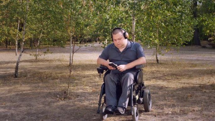 轮椅男子。残疾人。年轻的残疾人，坐在自动轮椅上，在秋天的晴天，在城市公园里，戴着耳机听音乐