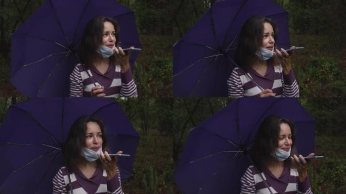 在阴天的秋天森林里，戴着冠状病毒面具的年轻白人妇女拿着雨伞，用智能手机。一名妇女在秋天的森林中的智能