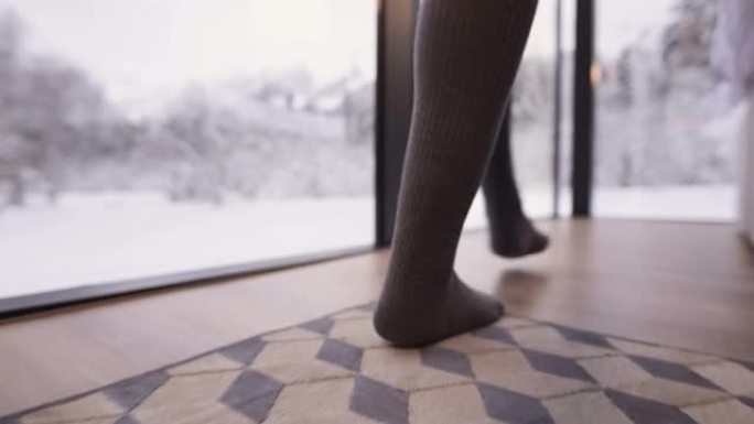一个特写镜头跟随一个穿着长统袜的女孩的脚，穿着袜子走过卧室里的一张床，卧室里有透明的墙壁窗户，后面是
